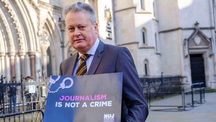 Vincent Kearney wearing a suit and tie holds a 'Journalism Is Not A Crime' placard.