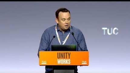 Fran McNulty wearing a blue shirt speaking behind a yellow lectern with the words 'Unity Works'.
