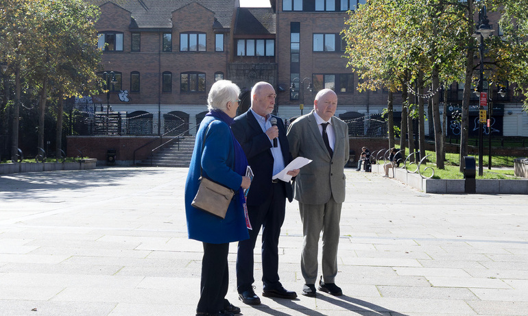 Kevin Cooper holding a written speech and a microphone alongside Anne Hailes and Séamus Dooley.