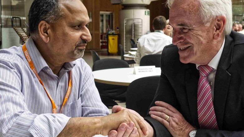 Barnie Choudhury and John McDonnell sitting at a table and shaking hands.