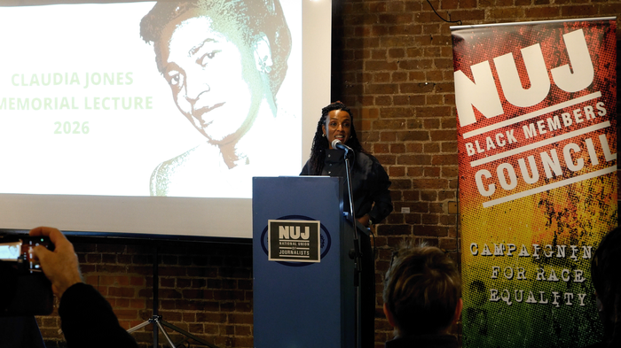 Nesrine Malik stands behind a blue podium with a projected image of Claudia Jones to her left and an NUJ Black Members' Council banner to her right.