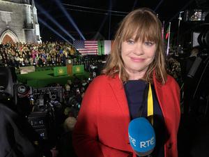 Teresa Mannion smiles as she holds a blue mic labelled RTE, whilst wearing a red coat.