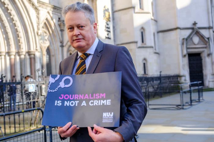 Vincent Kearney wearing a suit and tie holds a 'Journalism Is Not A Crime' placard.