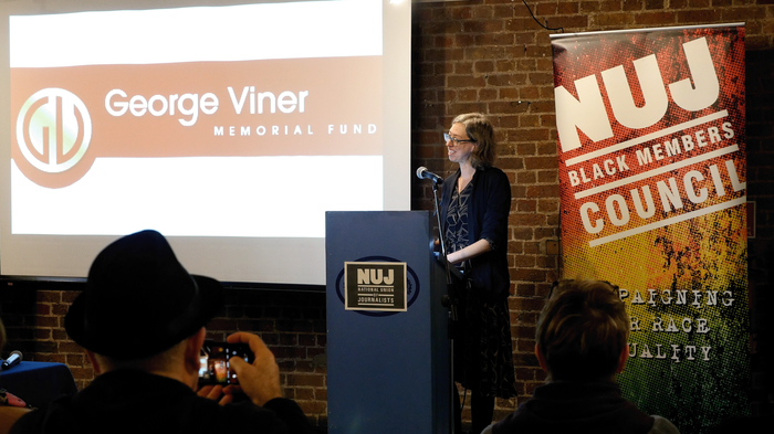 Laura Davison stands behind a blue podium with a projected image of Claudia Jones and two NUJ vertical banners behind her.
