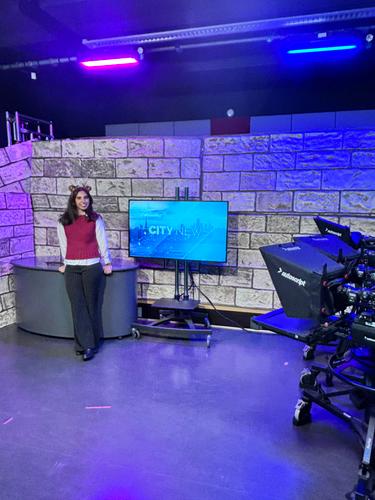 Hana Williams stands beside a television and desk in a studio with blue lighting.