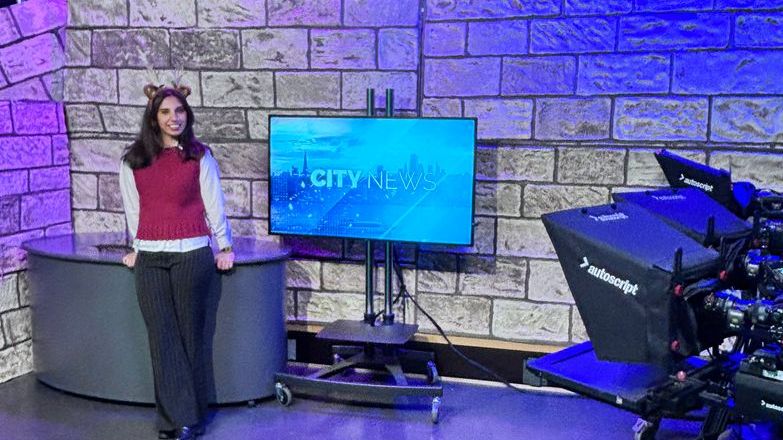 Hana Williams stands beside a television and desk in a studio with blue lighting.