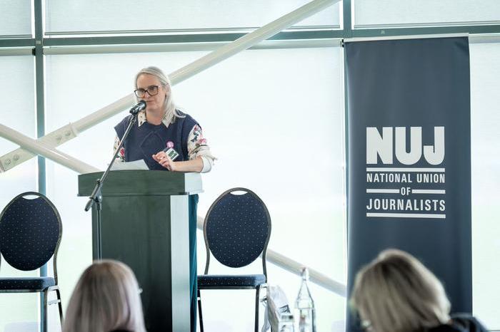 Rachel Howells speaking behind a lectern with a grey-green NUJ banner