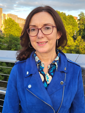 Máire T Ní Mhadaoin in blue jacket and dark spectacles smiles for a photo.