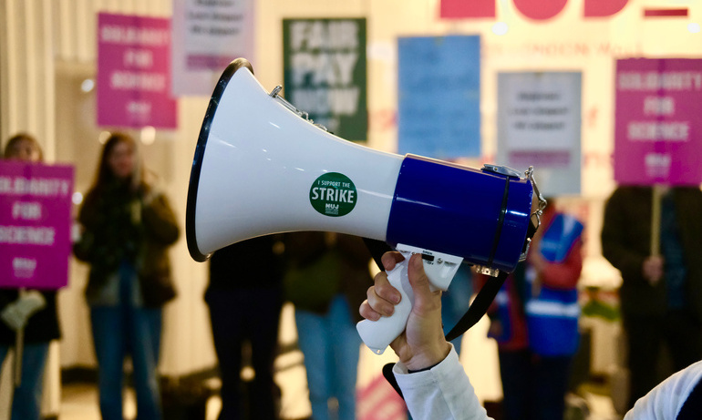 A white megaphone with a green NUJ sticker in the foreground with blurred green and pink placards in the background.