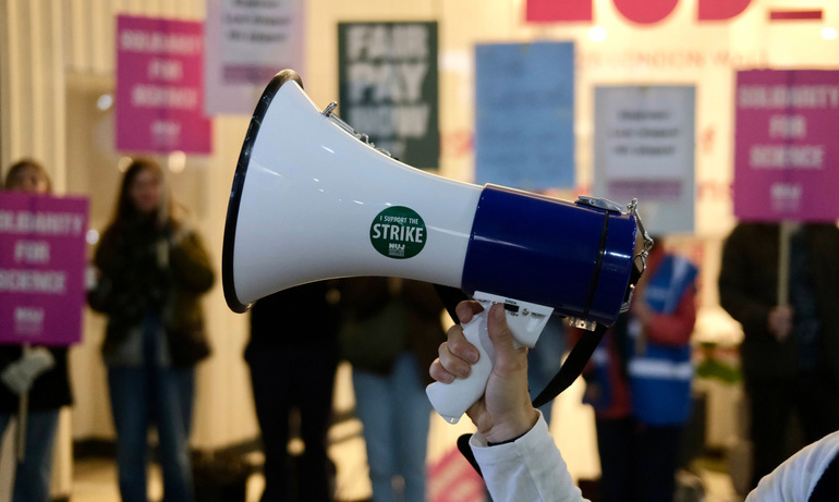 A white megaphone with a green NUJ sticker in the foreground with blurred green and pink placards in the background.
