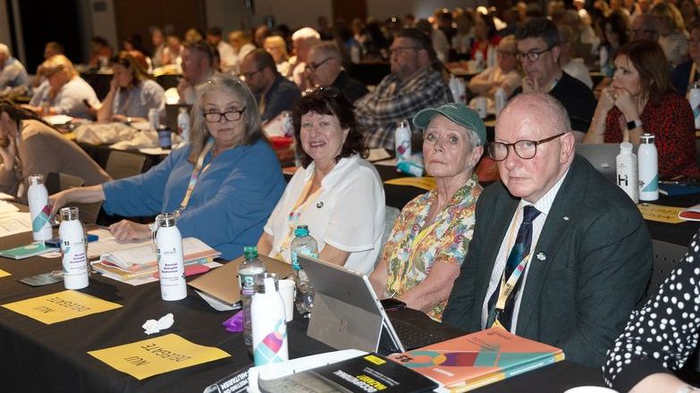 NUJ delegates sit at table with notepads and laptops as they pose for a photo. Others visible sitting at tables behind them. 