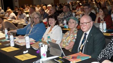 NUJ delegates sit at table with notepads and laptops as they pose for a photo. Others visible sitting at tables behind them. 