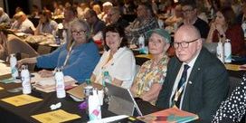NUJ delegates sit at table with notepads and laptops as they pose for a photo. Others visible sitting at tables behind them. 