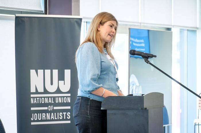 Laura Doel wearing a light blue top speaking behind a lectern and microphone with a dark-grey 'NUJ' banner in the background.