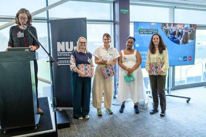 Laura Davison speaking on stage behind a lectern with Rachel Howells, Zena Chandler-Burnell, Huda Elmi and Natasha Hirst holding gift bags.