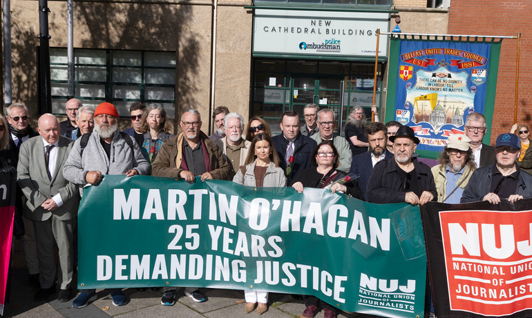 A crowd of people standing in a square holding a green banner that states 'Martin O'Hagan: 25 Years Demanding Justice' in front of a police building.