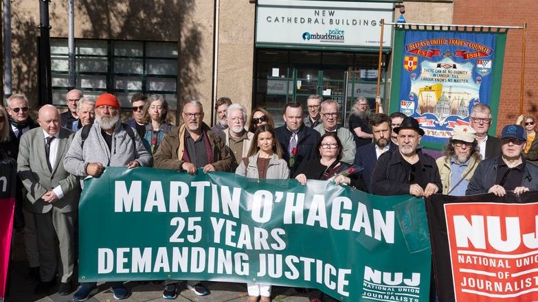 A crowd of people standing in a square holding a green banner that states 'Martin O'Hagan: 25 Years Demanding Justice' in front of a police building.