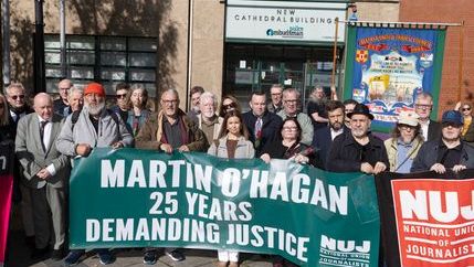 A crowd of people standing in a square holding a green banner that states 'Martin O'Hagan: 25 Years Demanding Justice' in front of a police building.