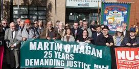 A crowd of people standing in a square holding a green banner that states 'Martin O'Hagan: 25 Years Demanding Justice' in front of a police building.