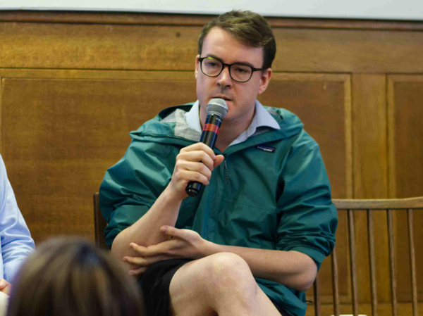 John McEvoy wearing a coat, short and glasses holding a microphone.