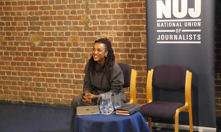 Nesrine Malik sitting behind a table and laughing with a green NUJ vertical banner to her right.