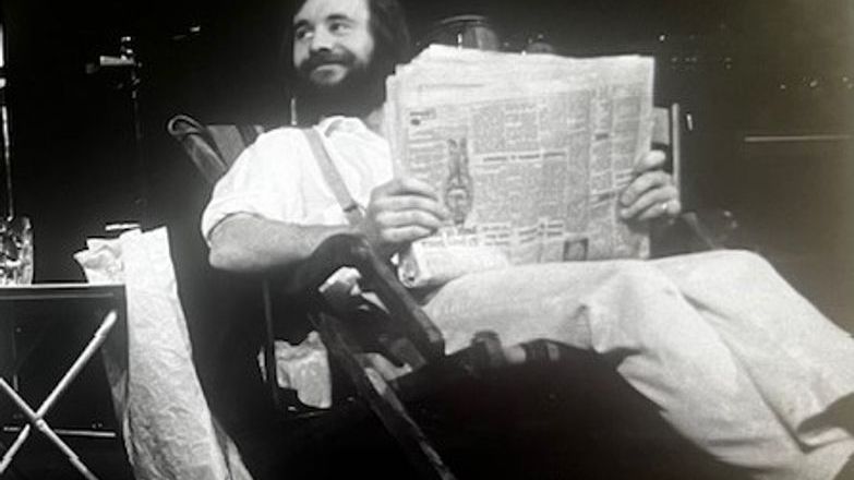 Black and white photo of John Mathews sitting in a chair and reading a broadsheet newspaper.
