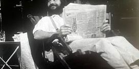 Black and white photo of John Mathews sitting in a chair and reading a broadsheet newspaper.
