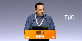 Fran McNulty wearing a blue shirt speaking behind a yellow lectern with the words 'Unity Works'.