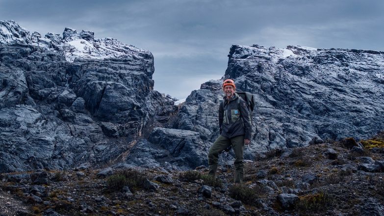 Klaus Thymann wearing hiking gear standing in front of mountainous, snow-topped rocks against a dark grey sky.