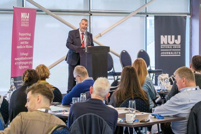 David Nicholson wearing a grey suit and tie speaking behind a lectern with a pink NUJ banner to his left and a dark green NUJ banner to his right.