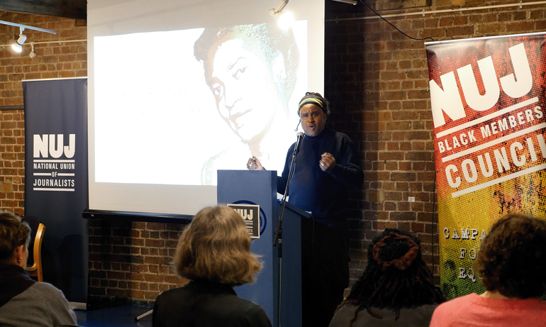 Roger McKenzie stands behind a blue podium with a projected image of Claudia Jones and two NUJ vertical banners behind him.