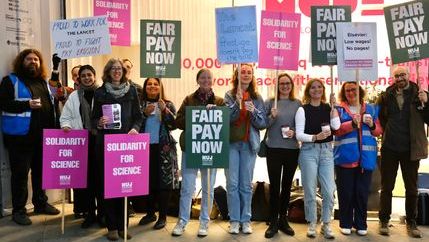 A group of people holding placards that say 'Fair Pay Now' and 'Solidarity for Science' with a glass building.
