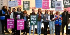 A group of people holding placards that say 'Fair Pay Now' and 'Solidarity for Science' with a glass building.