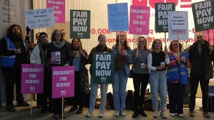 A group of people holding placards that say 'Fair Pay Now' and 'Solidarity for Science' with a glass building.