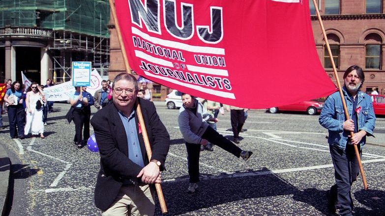 Martin O'Hagan wearing glasses and holding a red NUJ banner on a sunny day.