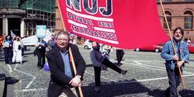 Martin O'Hagan wearing glasses and holding a red NUJ banner on a sunny day.