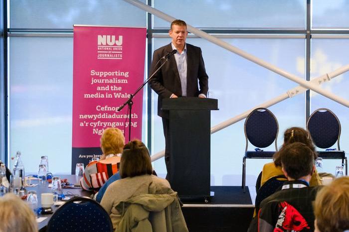Rhun ap Iorwerth wearing a suit standing behind a lectern with a pink NUJ banner to his left.