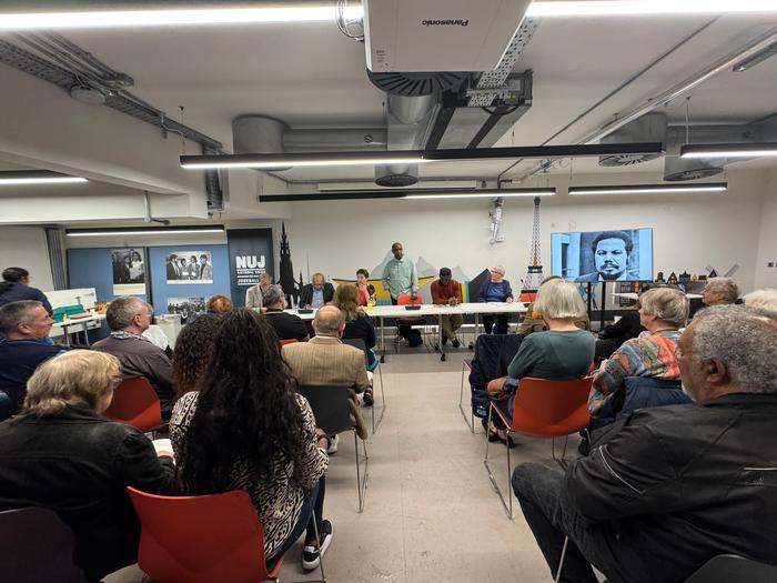 People seated in a basement with strip panel lightning and a table with a panel of speakers with images of Lionel Morrison on either side.