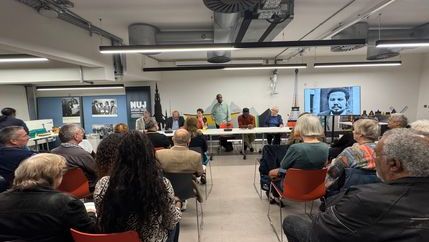People seated in a basement with strip panel lightning and a table with a panel of speakers with images of Lionel Morrison on either side.