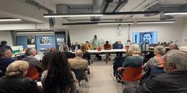 People seated in a basement with strip panel lightning and a table with a panel of speakers with images of Lionel Morrison on either side.