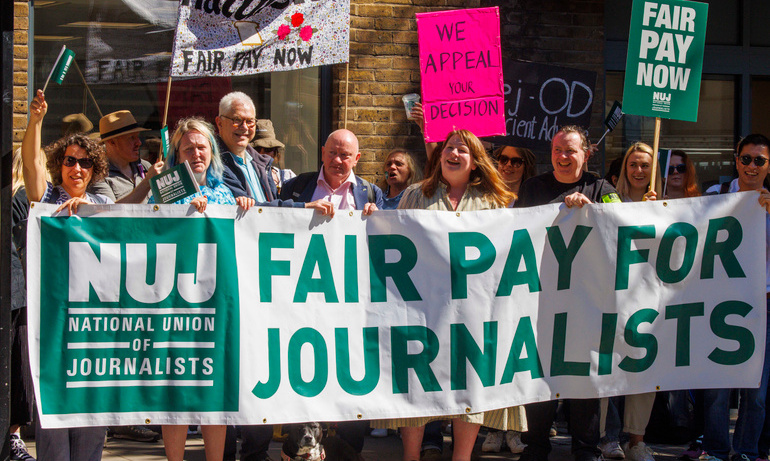 White banner with green text fair pay for journalists is held with placards visible in background
