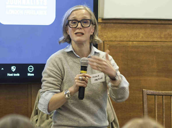Anna Hervé wearing glasses and a grey jumper holding a microphone.