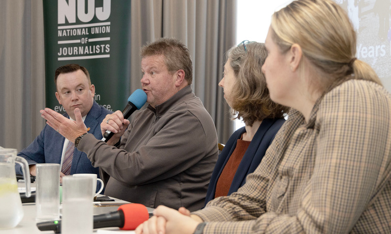 A man speaking into a microphone while three people seated either side of him look with attentive expressions.