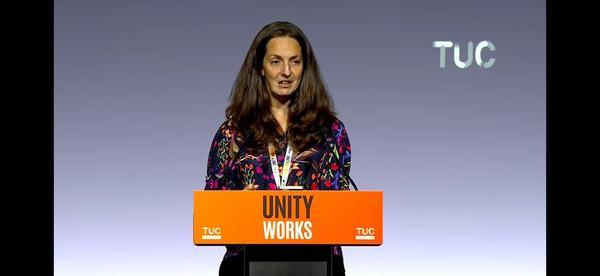 Natasha Hirst wearing a blue blouse speaking behind an orange lectern with the words 'Unity Works'.