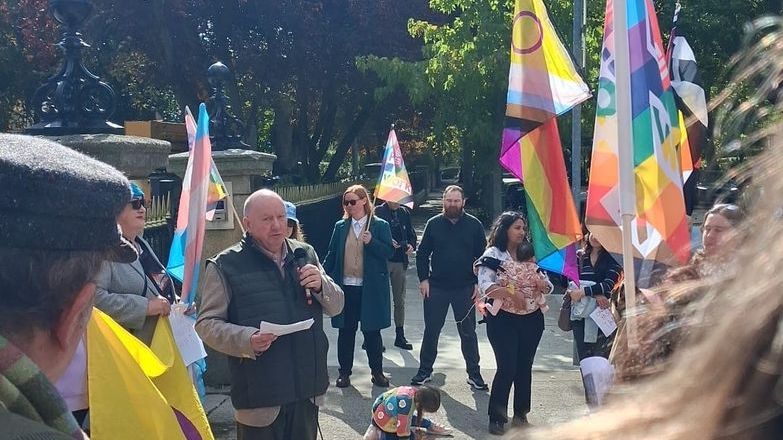 Séamus Dooley wearing a gilet and holding a microphone in a sunny square with colourful LGBT+ flags in the background.