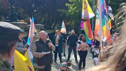 Séamus Dooley wearing a gilet and holding a microphone in a sunny square with colourful LGBT+ flags in the background.