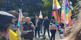 Séamus Dooley wearing a gilet and holding a microphone in a sunny square with colourful LGBT+ flags in the background.