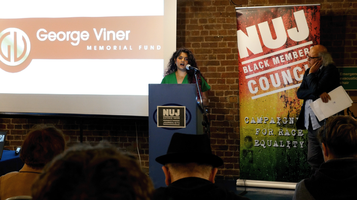 Meena Iyer stands behind a blue podium with a projected image of the GVMF logo to her left and an NUJ Black Members' Council banner to her right.
