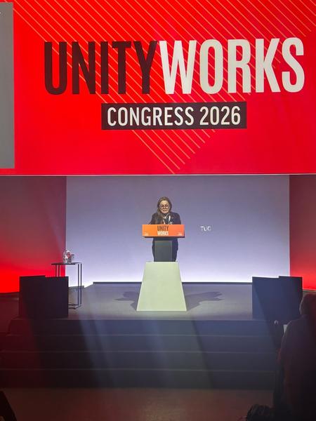 Pennie Quinton speaking at a lectern against a lilac background with a red banner above her with the words 'Unity Works'