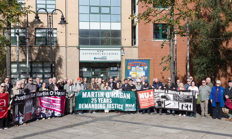 A crowd of people standing in a square holding a green banner that states 'Martin O'Hagan: 25 Years Demanding Justice' in front of a police building.